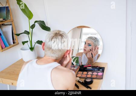 Uomo transgender che indossa un ombretto blu che applica mascara mentre guarda nello specchio Foto Stock