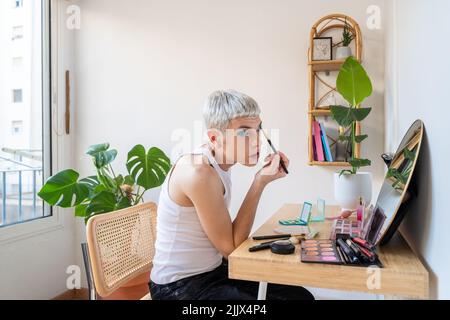 Uomo transgender che indossa un ombretto blu che applica mascara mentre guarda nello specchio Foto Stock