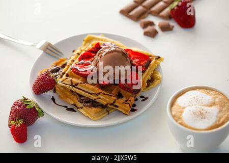 Da sopra gustosi waffle dolci fatti in casa al forno serviti su piatto con gelato alla fragola e cioccolato e salsa vicino tazza con caffè in kit leggero Foto Stock
