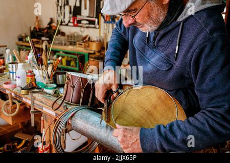 Artigiano maschio utilizzando le forbici mentre si tagliano i bordi metallici dei cuscinetti del tamburo mentre si lavora in officina professionale leggera Foto Stock