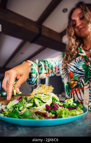 Attraente cuoco femminile mettendo cetriolo tritato in insalata fresca con germogli di fagiolo mungo in cucina leggera a casa Foto Stock