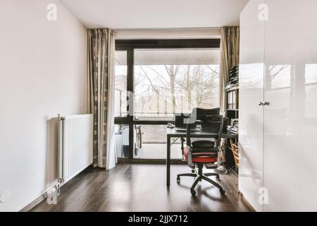 Tavolo con computer e decorazione situato vicino a grandi finestre e ripiani con documenti in un ufficio moderno e luminoso Foto Stock