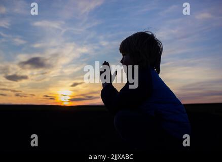 Silhouette di un ragazzo, ripiegò le mani in preghiera, girandosi verso Dio sullo sfondo di un pittoresco tramonto. Bambini contro la guerra. Bambino chiede pace f Foto Stock