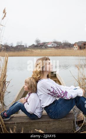 il ragazzino e una giovane bella donna in abiti nazionali ricamati si siedono di nuovo sulla riva del lago. Famiglia, rifugiati, unità, sup Foto Stock
