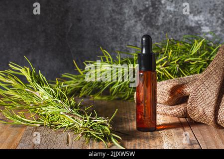 Su un tavolo di tavole di legno si trova una bottiglia di vetro marrone, con tappo contagocce, contenente olio essenziale circondato da ramificazioni di rosmarino e un sacco di stoffa. Foto Stock