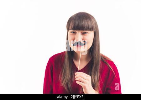 Giovane donna bianca con capelli scuri e bangs con maglione rosso che tiene fake baffi. Foto studio. Spazio di copia isolato. Foto di alta qualità Foto Stock