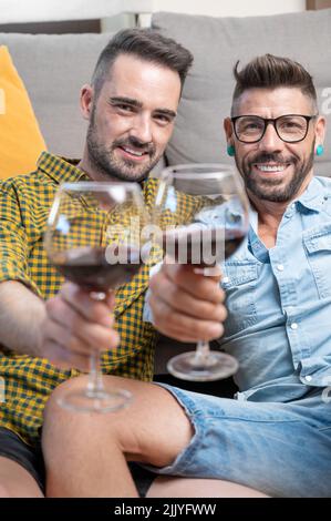 Coppia gay innamorata seduta a casa sul pavimento e tostatura con vino rosso. Fotografia di alta qualità. Foto Stock