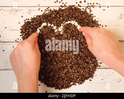 Mano femminile che disegna un simbolo di cuore nel mucchio di buckwheat organico buccia su un fondo di legno assi, usato come riempimento per cuscino di grano saraceno Foto Stock