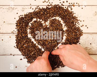 Mano femminile che disegna un simbolo di cuore nel mucchio di buckwheat organico buccia su un fondo di legno assi, usato come riempimento per cuscino di grano saraceno Foto Stock