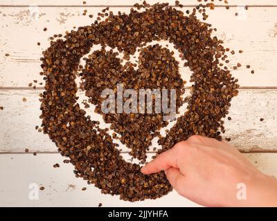 Mano femminile che disegna un simbolo di cuore nel mucchio di buckwheat organico buccia su un fondo di legno assi, usato come riempimento per cuscino di grano saraceno Foto Stock