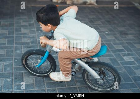 Ragazzo caucasico che gioca con la bicicletta nel cortile della sua casa. Ragazzo a cavallo veloce intorno casa privata. Giochi per bambini e sport ciclistici. Foto Stock