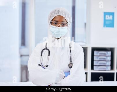 Il suo tempo di affrontare il covid. Ritratto corto di un medico fidato in piedi con le braccia ripiegate mentre vestito in tute PPE in ufficio. Foto Stock