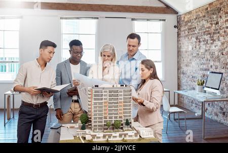 Un team diversificato di architetti che lavorano su un modello di costruzione insieme all'interno dell'ufficio. Un gruppo di ingegneri che pianificano un progetto di ricostruzione o progettazione Foto Stock