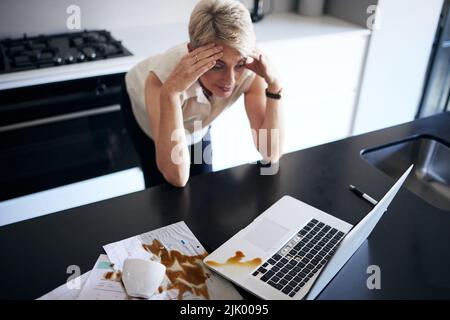 Non so cosa fare. Una donna matura si sente stressata dopo aver sparso il caffè sulla sua carta e computer portatile in cucina a casa. Foto Stock