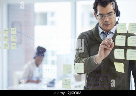 Un giovane uomo d'affari che indossa una cuffia mentre si fa il brainstorming con note su una parete di vetro in un ufficio. Foto Stock