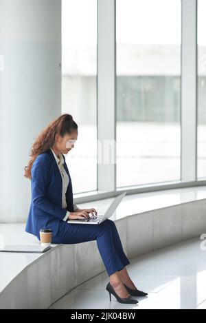 Giovane donna vietnamita che siede alla grande finestra dell'ufficio e lavora sul computer portatile Foto Stock