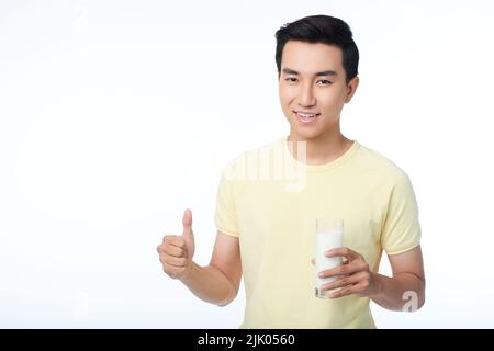 Giovane uomo fiducioso con un sorriso toothy che mostra il pollice in su e tiene in mano un bicchiere di latte mentre si alza su sfondo bianco, ritratto vita-su colpo Foto Stock