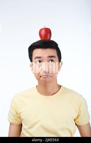 Uomo asiatico pensivo guardando verso l'alto mentre bilanciamento mela rossa sulla sua testa, isolato su sfondo bianco Foto Stock