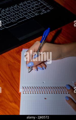 Giovane donna che lavora da casa con un computer portatile mentre prende appunti in un notebook. Foto Stock