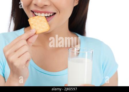 Primo piano di una giovane donna irriconoscibile che mangia il cracker e beve il latte mentre si alza su sfondo bianco Foto Stock