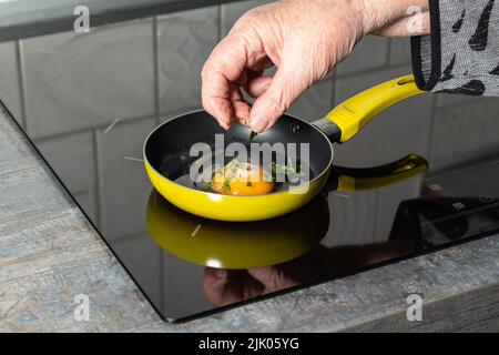 La mano della donna anziana irrora l'uovo crudo con sale e erbe tritate, fuoco selettivo. Primo piano di piastra in ceramica, piano in legno e bellissimo giallo f Foto Stock