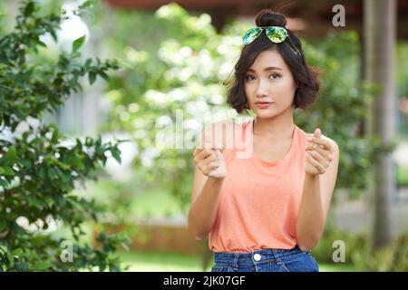 Ritratto di attraente donna asiatica con taglio di capelli alla moda guardando la macchina fotografica mentre balla al suo brano preferito, cortile verde illuminato con raggi del sole sullo sfondo Foto Stock
