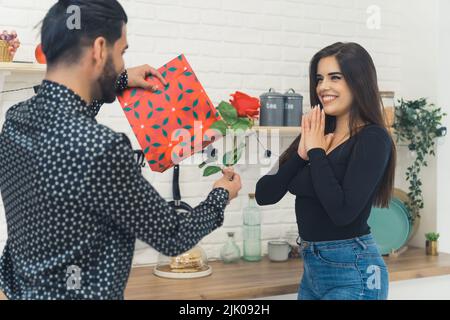 L'uomo che indossa una camicia a motivi geometrici dà il regalo di giorno della sua ragazza San Valentino e rosa in cucina. Donna sorpresa sorridendo larga. Scatto al coperto. Foto di alta qualità Foto Stock