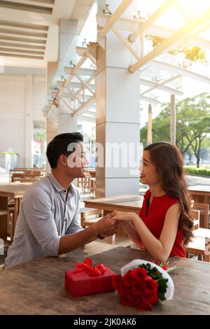 Adorabile coppia asiatica che si guarda l'un l'altro con sorrisi ampi e le mani in mano mentre festeggia il giorno di San Valentino al bar all'aperto, bouquet di rose rosse e scatola regalo che si trova sul tavolo Foto Stock