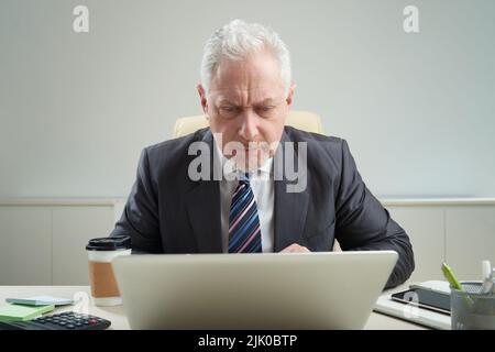 Ritratto in vita di uomo d'affari anziano concentrato indossando abito classico seduto alla scrivania d'ufficio e lavorando su un importante progetto con l'aiuto di un laptop Foto Stock
