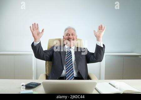 Un manager gioioso e anziano in abbigliamento formale seduto alla scrivania con le mani sollevate e guardando la fotocamera con un sorriso toothy, l'interno dell'ufficio sullo sfondo Foto Stock