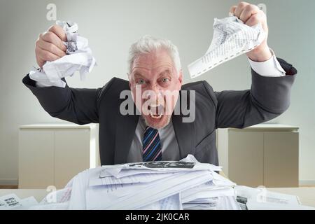 MAD uomo d'affari anziano indossando abito classico che esprime la sua sensazione negativa mentre si siede alla scrivania d'ufficio: Gridando e sgretolando documenti, ritratto girato Foto Stock