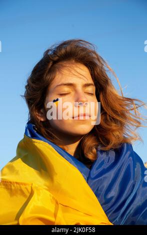 ritratto di una giovane bella donna stanca ma non rotta con un cuore dipinto giallo-blu sulla guancia e avvolto in una bandiera. Luce del sole al tramonto. Supporto Foto Stock