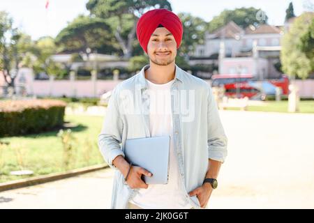 Lavorare con gioia. Sorridente uomo libero indiano allegro indossando tradizionale Headwrap turban in piedi con computer portatile all'aperto, guardando la fotocamera, imprenditore di successo maschile Foto Stock