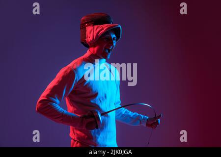 Ritratto di giovane fencer maschio alto in costume da scherma e maschera che tiene spada piccola isolato su sfondo viola in neon. Sport, emozioni, energia Foto Stock