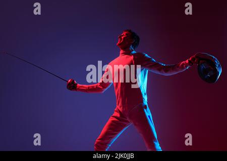 Ritratto di giovane fencer maschio alto in costume da scherma e maschera che tiene spada piccola isolato su sfondo viola in neon. Sport, emozioni, energia Foto Stock