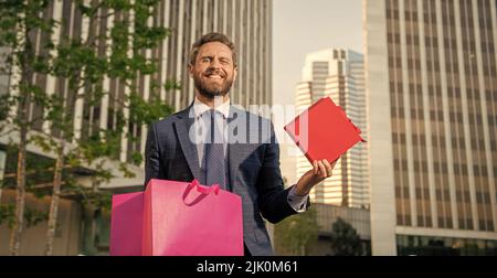 felice uomo riuscito in formalwear messo scatola presente in borsa della spesa fuori dell'ufficio, shopping Foto Stock
