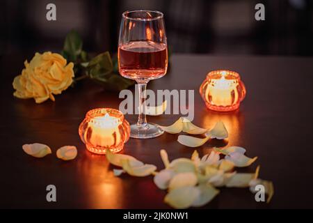 Un bicchiere di vino di rosa, due candele e petali di rosa su un tavolo scuro Foto Stock