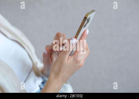 A close-up photo of a beautiful woman's hand with a white manicure is using a gold-colored phone, typing a message on a smartphone Foto Stock