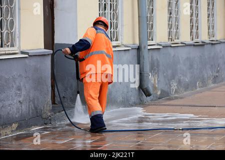 L'operatore innaffia il marciapiede con un tubo flessibile. Pulizia e disinfezione della strada nella città estiva Foto Stock