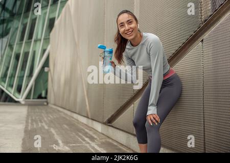 Fitness donna in sportswear bere acqua da una bottiglia dopo l'allenamento appoggiata sul muro Foto Stock