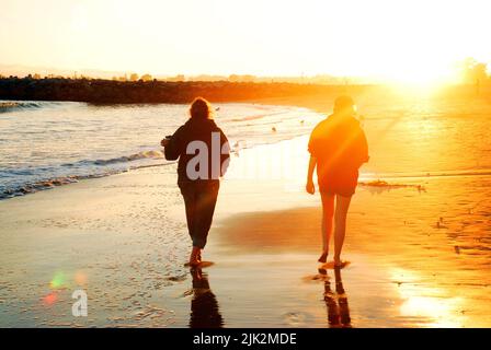 Due amici camminano lungo la spiaggia e il bordo dell'acqua al tramonto Foto Stock