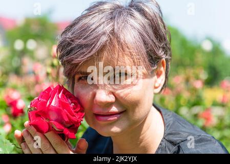 Primo piano di una donna leggermente sorridente che gode il profumo di una rosa rossa su uno sfondo sfocato di fiori in una giornata di sole. Foto Stock