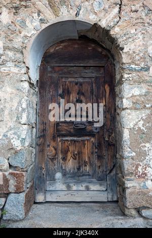 Vecchia porta in legno intemperie con piastre di bloccaggio arrugginite e maniglia in un muro, mascherata con grandi pietre naturali Foto Stock