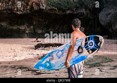 Vista posteriore di professionista surfista nero tenendo la sua tavola da surf in mano si alza su una spiaggia oceano contro la scogliera guardando la giovane donna prendere il sole Foto Stock