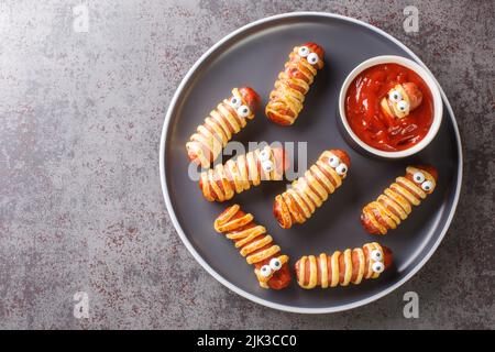 Divertenti mummie di salsiccia in pasta con ketchup per la festa di Halloween. Cibo per bambini in primo piano nel piatto sul tavolo. Vista dall'alto orizzontale Foto Stock