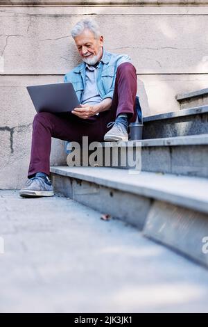 Un uomo anziano controlla le sue e-mail sul computer portatile mentre si siede sulle scale del parco. Foto Stock