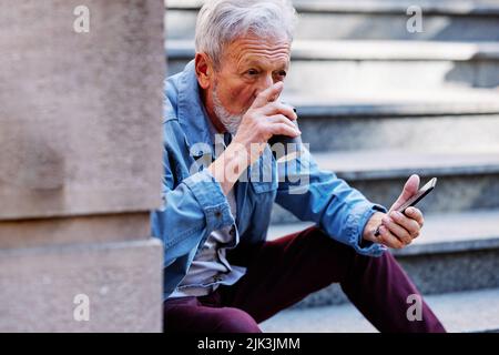 Un uomo anziano seduto sulle scale fuori, bevendo caffè e aspettando messaggi al telefono. Foto Stock