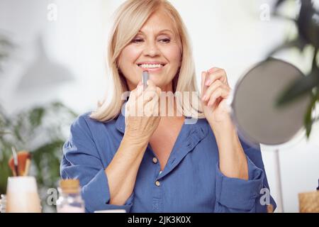 Una bella donna bionda anziana seduta a casa e mettere il rossetto sulle labbra. Foto Stock
