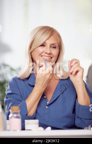 Una bella donna bionda anziana seduta a casa e mettere il rossetto sulle labbra. Foto Stock