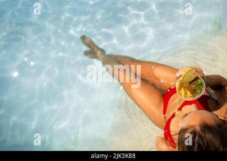 Donna in bikini rosso con un mojito cocktail all'interno della piscina in estate, vista aerea. Vacanze e concetto gioioso Foto Stock